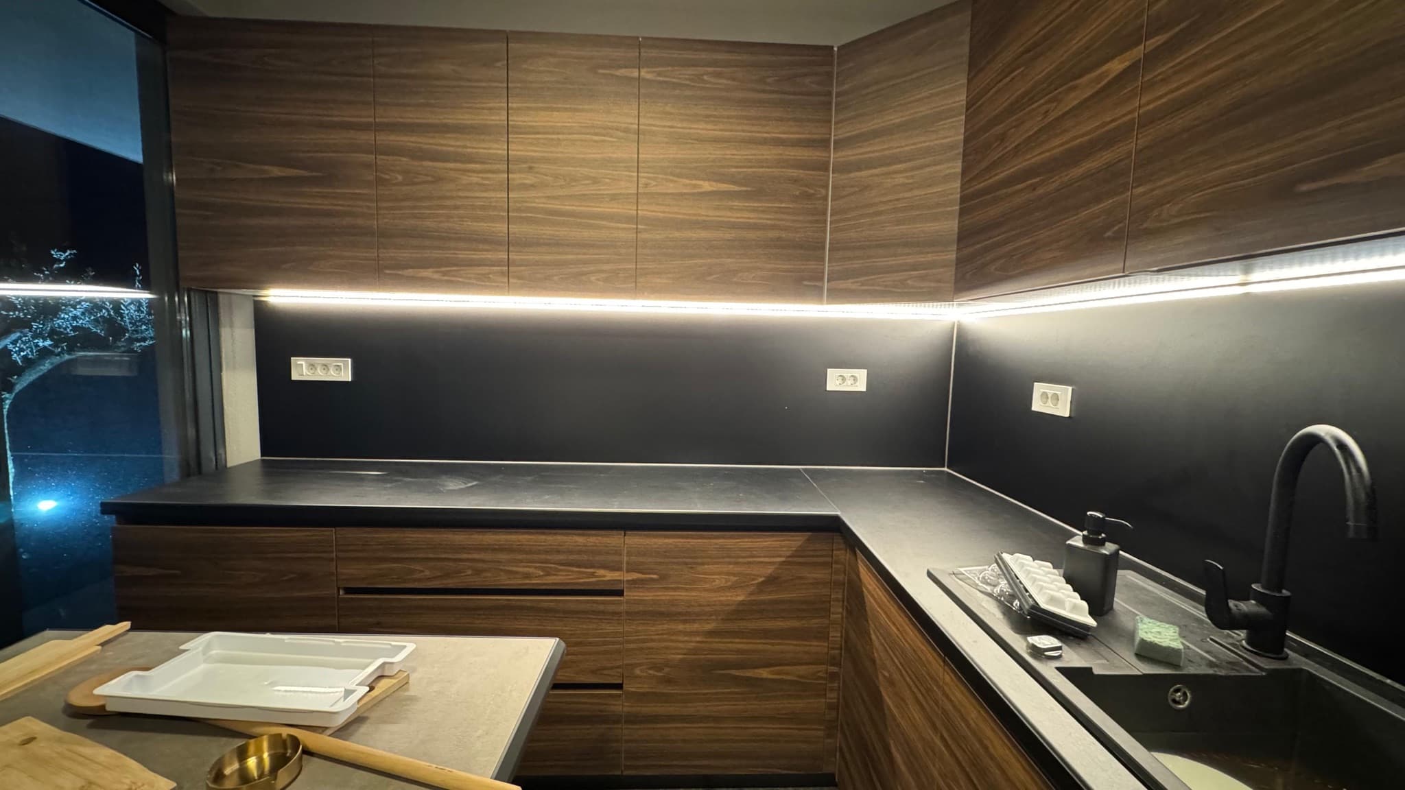 LED strip under the wall units lighting the worktop of a dark-veneer kitchen.
